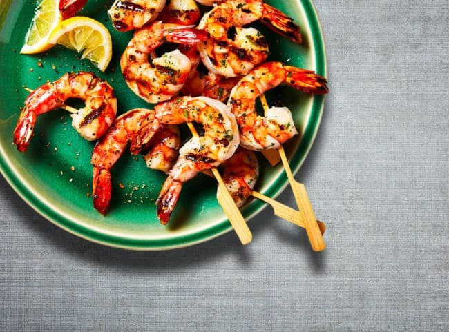 Round green plate with three cooked shrimp skewers laid on top, and cut limes for garnish